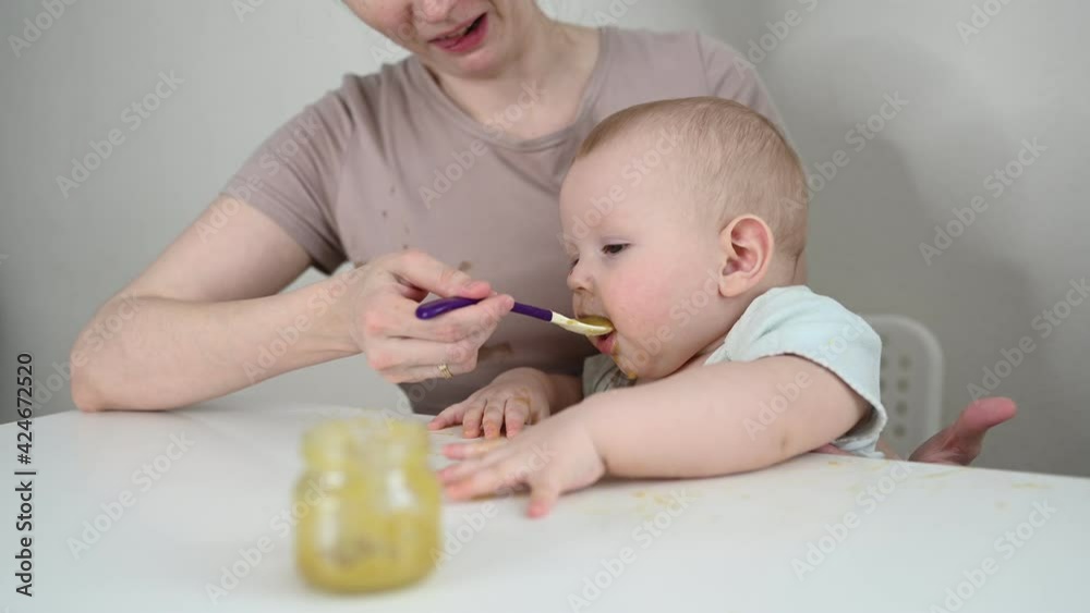 Little newborn funny baby boy learning to eat vegetable or fruit puree from glass jar with spoon. Young mother helping little son eat first food Little newborn funny baby boy learning to eat vegetable or fruit puree from glass jar with spoon. Young mother helping little son eat first food