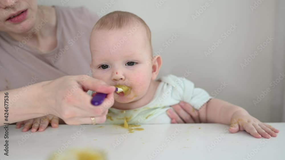 Little newborn funny baby boy learning to eat vegetable or fruit puree from glass jar with spoon. Young mother helping little son eat first food Little newborn funny baby boy learning to eat vegetable or fruit puree from glass jar with spoon. Young mother helping little son eat first food