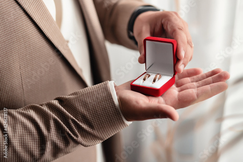 The man the groom in a white shirt and brown jacket holds on his palm wedding gold rings in a red velvet box by the window. Groom morning, close-up without a face only hands