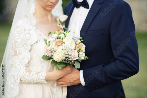 The groom in a blue suit, black leather shoes, socks with hearts and a bride in a white silk dress are sitting on a bench and holding a wedding bouquet together. Horizontal photo.