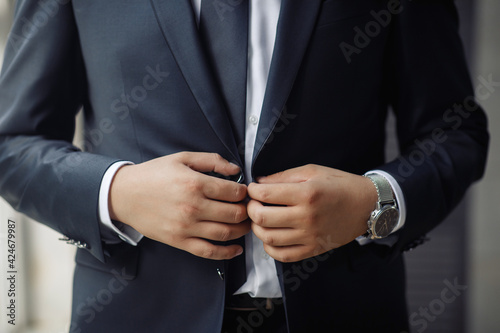 A man, a groom in a white shirt and black stylish checkered suit with a large stylish watch, fastens his jacket near the window. Morning groom, a guy without a face.