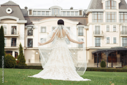 Girl brunette bride in a white dress with lace holds a long veil with her hands and looks at a large vintage house with a tower on the background of her back to the camera. Wedding day  walk in nature