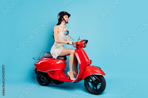 Full size profile side photo of young attractive positive smiling girl in helmet riding moped isolated on blue color background
