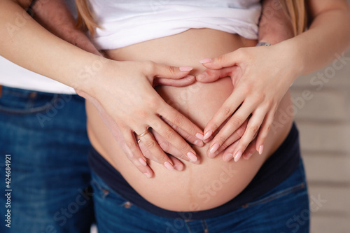 A man and a woman in white T-shirts and jeans on a black background are holding their hands in the shape of a heart on a bare pregnant belly. Loving couple awaiting the birth of a baby