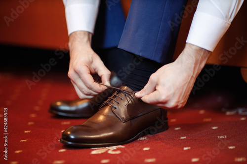 A strong man with his arms folded in a suit is tying shoe laces on brown leather classic shoes. A groom with a ring on his hand in shoes for his wedding. Only hands without a face.