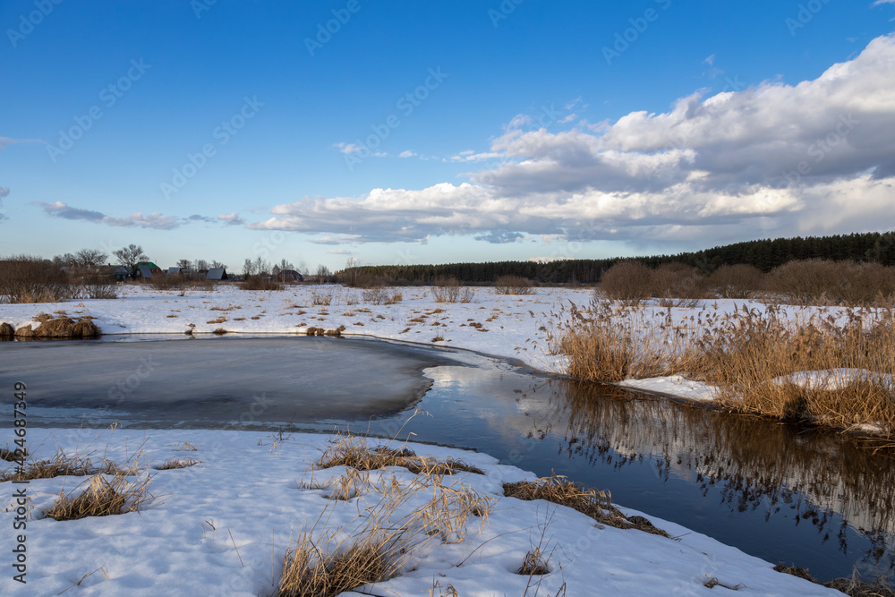 Early spring, blue sky and clouds are reflected in the river. Grass and snow in the foreground. Ice is melting on the river.