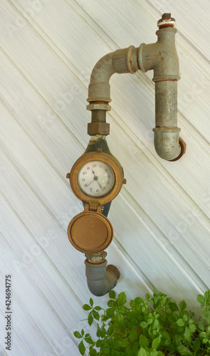 Water clock with classic pointer of an analog clock hangs with piping on a white wood paneling. Green shrub in the corner. Germany. photo