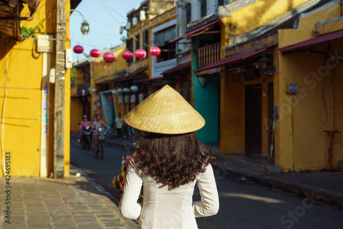 A young girl in traditional dress Ao Dai and conical hat standing on Hoi An ancient street