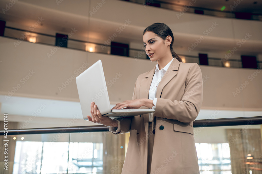Young businesswoman in beige jacket standing with a laptop and looking concentrated Young businesswoman in beige jacket standing with a laptop and looking concentrated