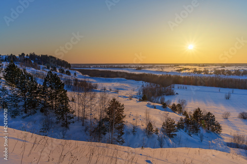 Sunset over the Irtysh river valley in Siberia (Russia) on a cold winter evening. Bird's eye view of the frozen river. There are many trees and vegetation along the banks. Blue-yellow sky. Rainbow sun © olgaS
