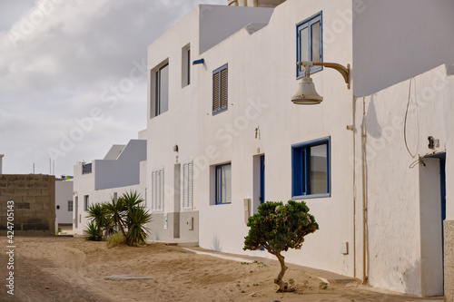 calle de arena, a la derecha casas con fachada blanca y ventanas azules con unos arboles en la puerta. Isla La Graciosa, Canarias, Spain