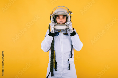 Male cosmonaut in spacesuit and helmet, holding his helmet with his hands with surprise gesture, on yellow background.