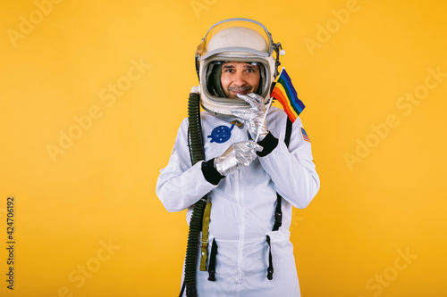 Murais de parede Gay male cosmonaut in space suit and helmet, holding lgtbi rainbow flag, on yellow background