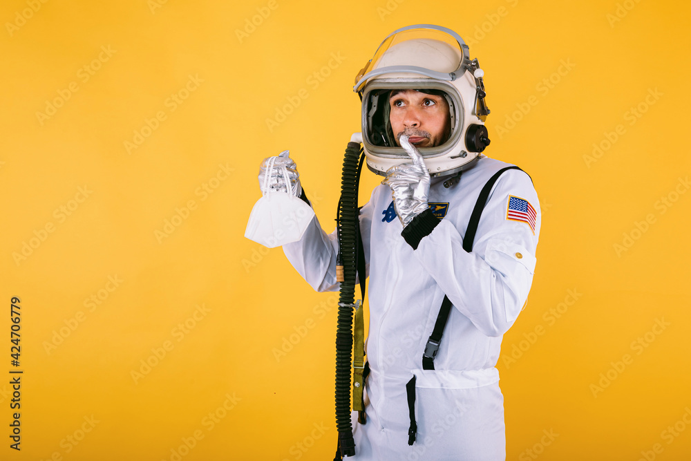 Male cosmonaut in spacesuit and helmet, holding a FPP2 mask, on yellow background. Covid-19 and virus concept Male cosmonaut in spacesuit and helmet, holding a FPP2 mask, on yellow background. Covid-19 and virus concept