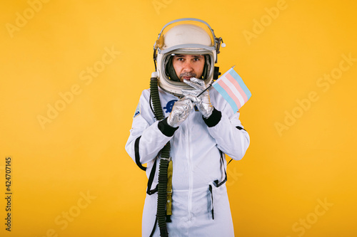 Slika na platnu Gay transsexual male cosmonaut with serious gesture in space suit and helmet, holding transgender flag, on yellow background