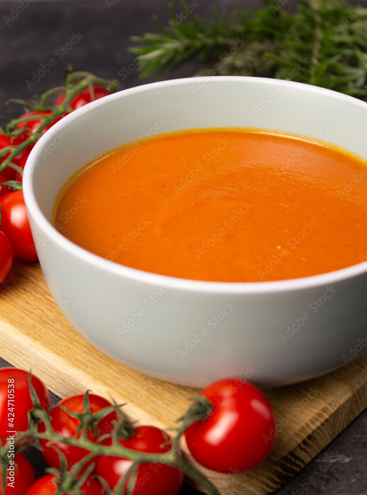 Rich color tomato puree soup in a gray bowl on a wooden board with cherry tomatoes and herbs on a background