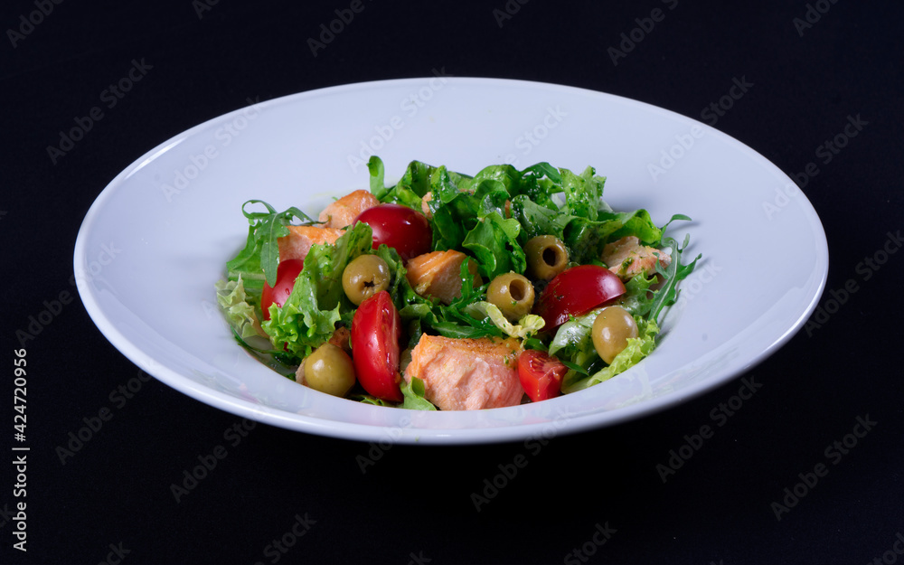 Fresh salmon salad, cherry tomatoes, olives, green lettuce, arugula in a white plate on a black background