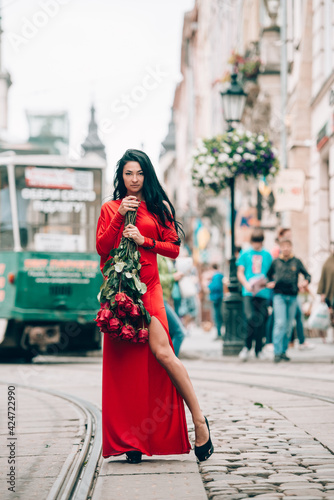 Charming young woman in red sexy dress posing with a bouquet of red roses. photo of a seductive woman with black hair on the tram track. Selective focus, filmgrain