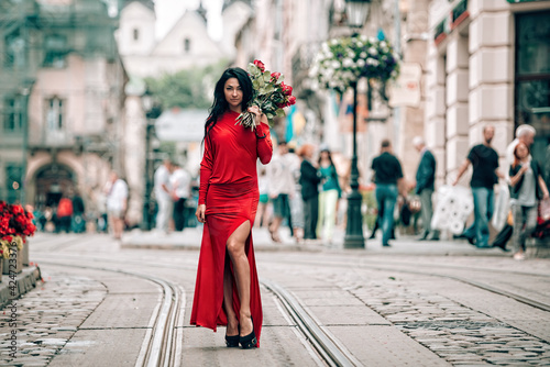 Charming young woman in red sexy dress posing with a bouquet of red roses. photo of a seductive woman with black hair on the tram track. Selective focus, filmgrain