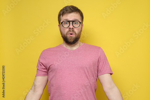 Surprised bearded man with glasses looks at the camera with big eyes and open mouth. Yellow background.