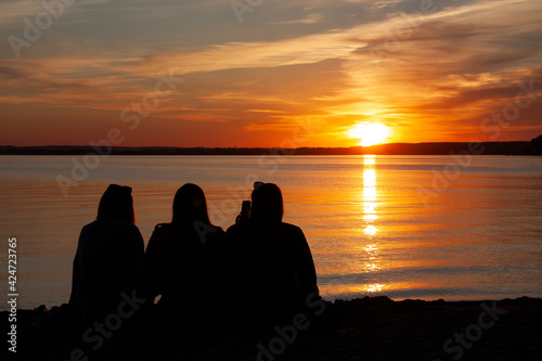 Horizontal conceptual romantic photo of three friends silhouettes with their back to the camera, sitting and relaxing on the beach during calm orange sunset over lake