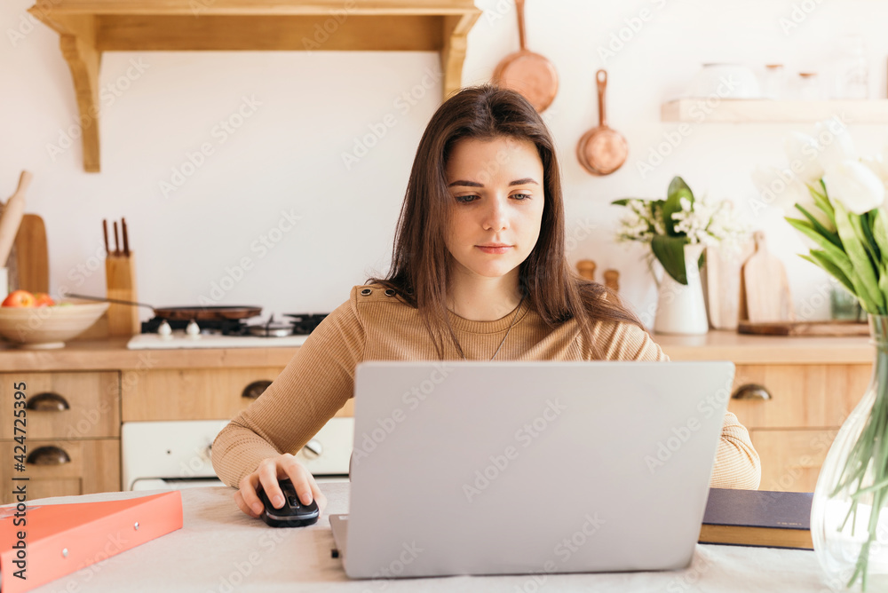 Teen girl with laptop doing homework in bright kitchen with flowers on the table. education and business concept. remote work and freelance work. student. Teen girl with laptop doing homework in bright kitchen with flowers on the table. education and business concept. remote work and freelance work. student.