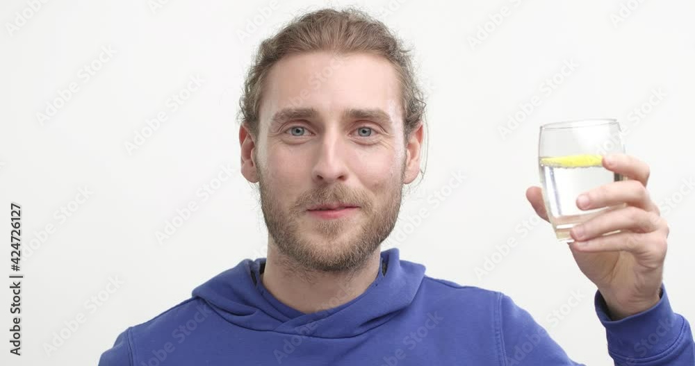 Happy bearded Young Man Drinking water with lemon from glass on White Background. Showing Healthy Lifestyle. Standing at studio wearing Blue Sweatshirt. Having Healthy Skin. Life. Water Balance. Happy bearded Young Man Drinking water with lemon from glass on White Background. Showing Healthy Lifestyle. Standing at studio wearing Blue Sweatshirt. Having Healthy Skin. Life. Water Balance.