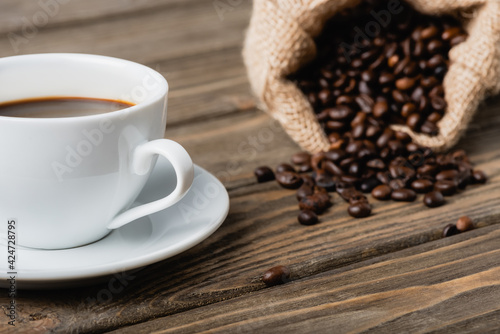 blurred sack bag with roasted coffee beans near cup on wooden surface