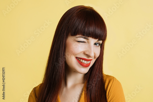 People, emotions, facial expressions. Close up portrait of jolly, vivacious sttractive young woman long straight red hair, winking to camera with smile, on isolated yellow background