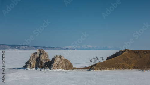 The frozen lake is covered with ice and snow. A picturesque two-headed rock rises above its surface. Tiny figures of people walking are visible. A mountain range in the distance. Clear sky. Baikal