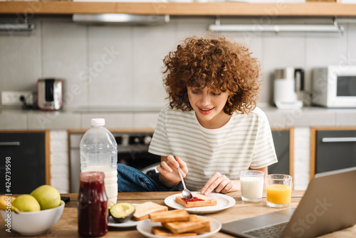 Smiling young woman using laptop while having breakfast at home kitchen