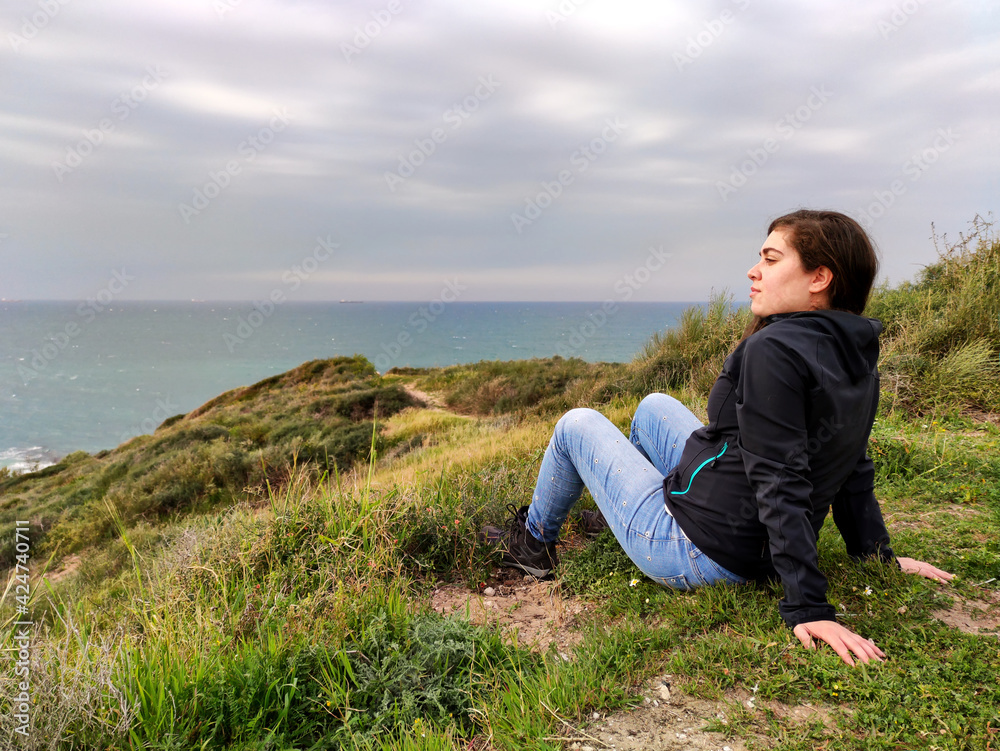 Young woman sitting on some rocks, on top of some dunes, watching the sea.