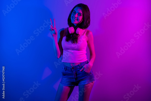 Photo of young smiling positive beautiful girl showing v-sign wear headphones isolated on blue glowing vibrant background
