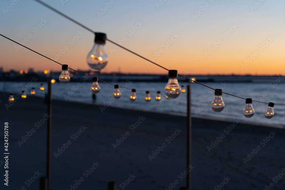 Close up of the glowing light bulbs at the beach cafe terrace. Sun is setting in the blurry abstract background.
