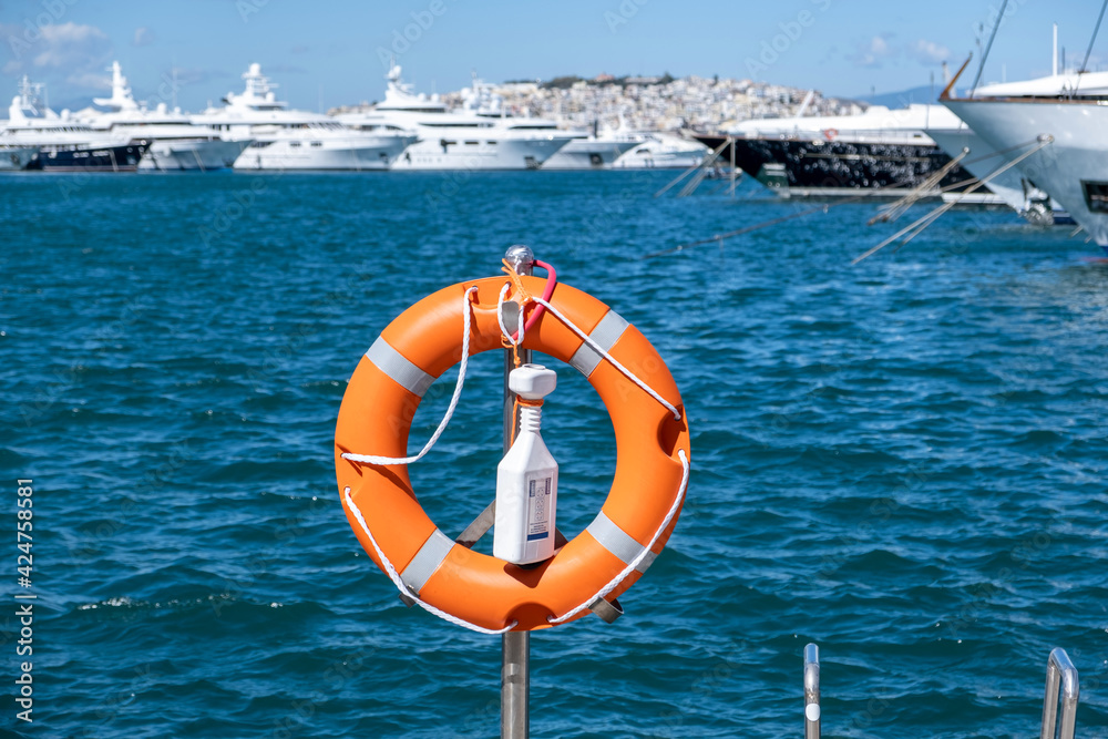 Lifebuoy orange on vertical iron, sea, boats, town background.