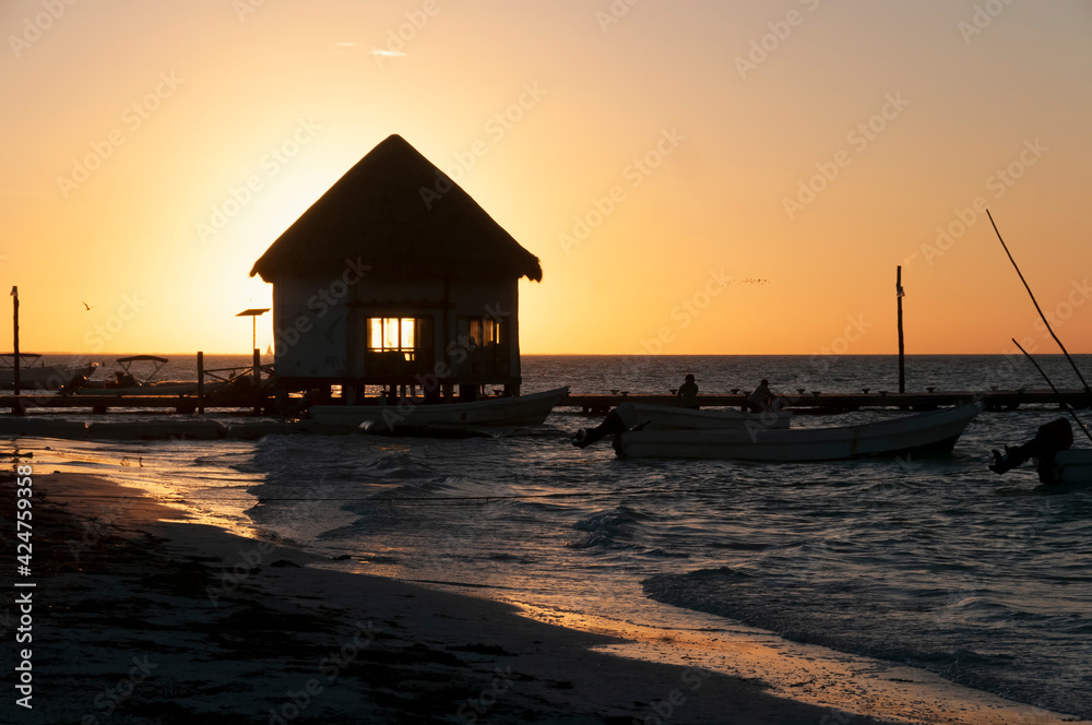 A small fishing shack on a wooden pier over the sea at sunset on Holbox Island in Mexico. In the background the sky and fishing boats. Travel Tourism Concept A small fishing shack on a wooden pier over the sea at sunset on Holbox Island in Mexico. In the background the sky and fishing boats. Travel Tourism Concept