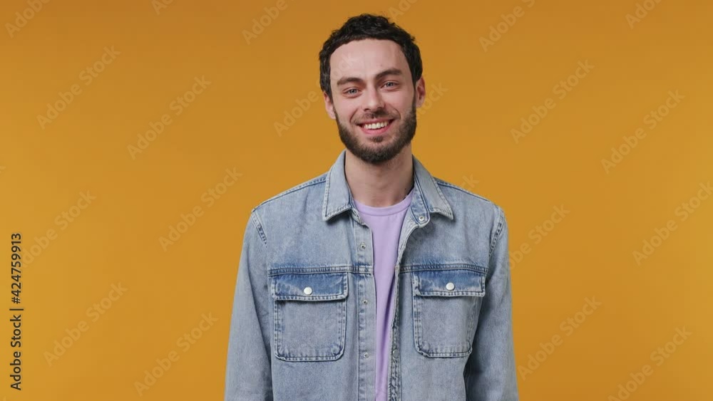 Handsome unshaven bearded young man 20s in denim jacket purple t-shirt looking camera asides charming smile isolated on yellow color wall background studio portrait People emotions lifestyle concept