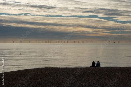 A couple sit on Brighton Beach looking out at Rampion Wind Farm on the horizon. photo