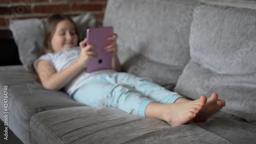 A young girl lies at home on the couch and looks into a pink tablet. She is funny and emotional. The child is dressed in pajamas and has bare feet.  Focus on the feet. 4K