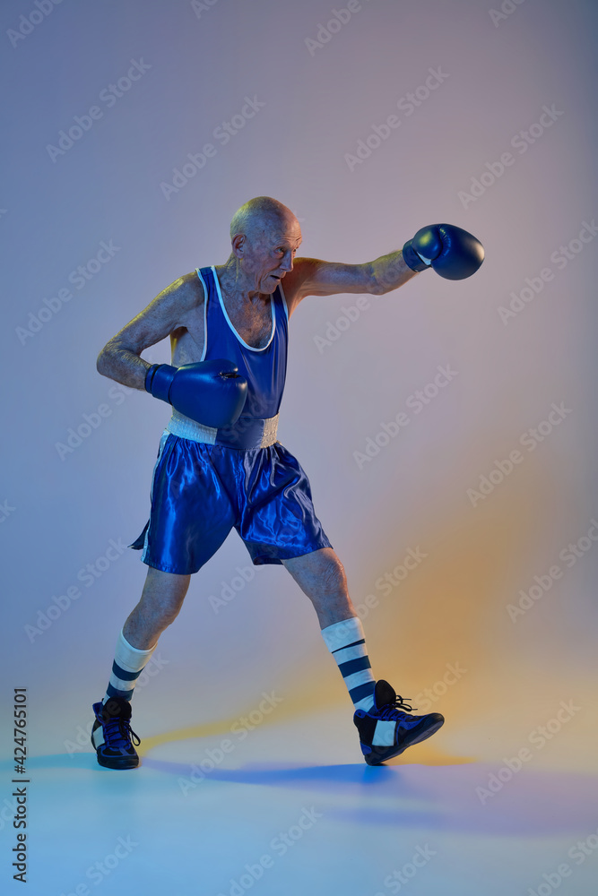 Senior man wearing sportwear boxing isolated on gradient studio background in neon light. Concept of sport, activity, movement, wellbeing. Copyspace, ad.