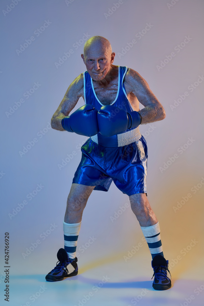 Senior man wearing sportwear boxing isolated on gradient studio background in neon light. Concept of sport, activity, movement, wellbeing. Copyspace, ad. Senior man wearing sportwear boxing isolated on gradient studio background in neon light. Concept of sport, activity, movement, wellbeing. Copyspace, ad.