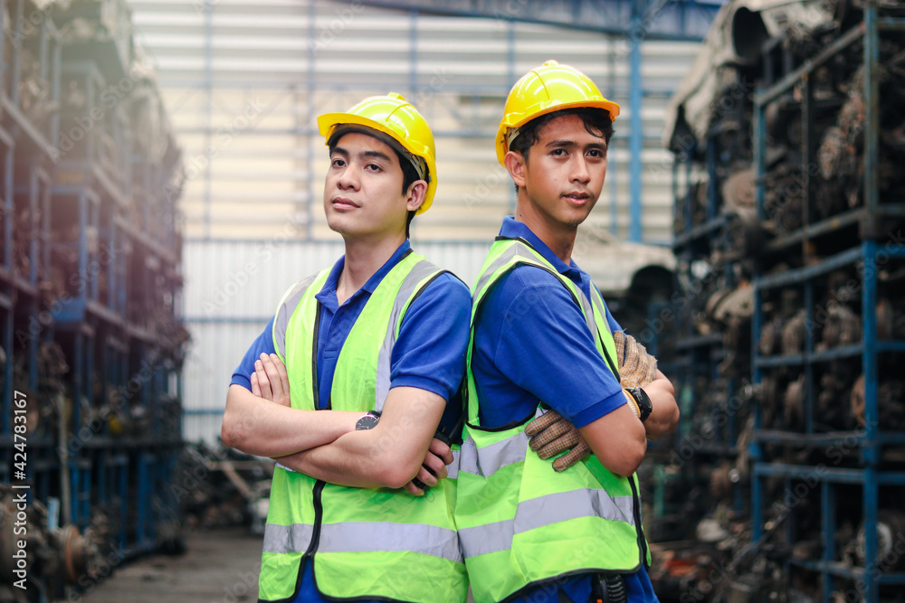 Portrait of two industrial engineer workers man wearing helmet with arms crossed, standing at manufacturing plant factory with many engine parts as blurred background Portrait of two industrial engineer workers man wearing helmet with arms crossed, standing at manufacturing plant factory with many engine parts as blurred background