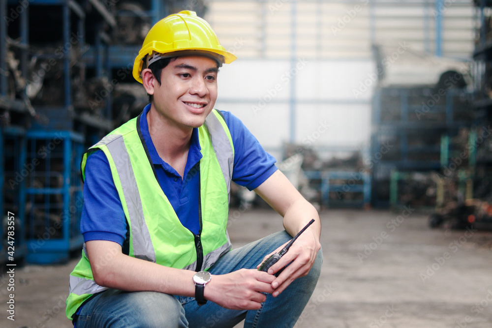 Industrial smiling worker wearing helmet holding radio walkie talkie at manufacturing plant factory industry, Asian young male engineer with many engine parts as blurred background Industrial smiling worker wearing helmet holding radio walkie talkie at manufacturing plant factory industry, Asian young male engineer with many engine parts as blurred background