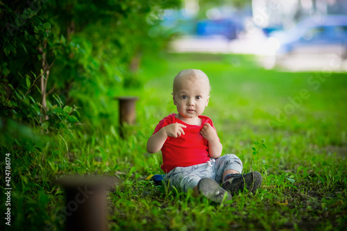 A 2-year-old kid in a bright red T-shirt on a background of green grass. Happy children in the summer on the street. Outdoor games and entertainment.