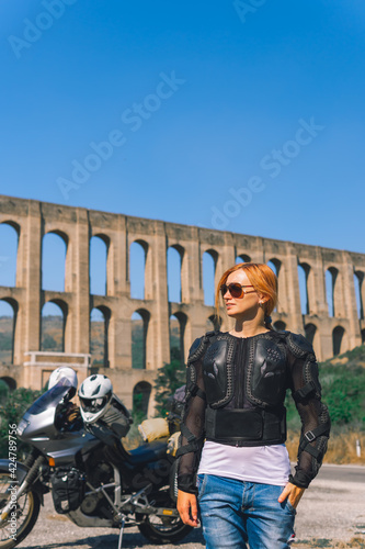 A girl in a protective motorcycle outfit poses against the background of an old Aqueduct of Vanvitelli, Caroline. Sunny day. Glasses and turtle. Motorbikers and travel. Vertical photo photo