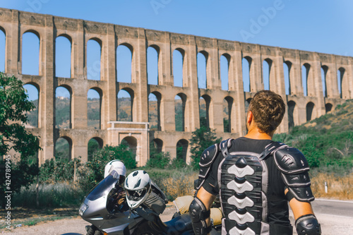 A man in a protective motorcycle outfit poses against the backgrond of an old Aqueduct of Vanvitelli, Caroline. Sunny day. Glasses and turtle jacket. Motorbikers and travel. photo