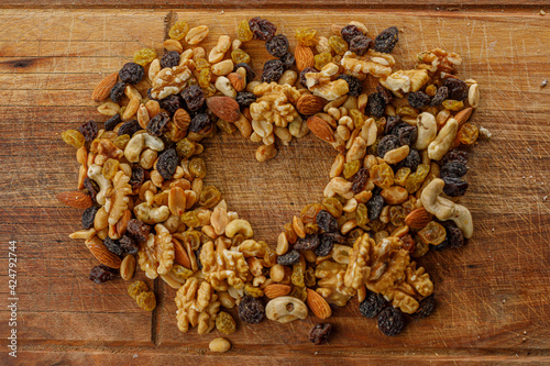 A top view of mix nuts. Raisins and a variety of dried fruits, grouped together forming a heart on a wood texture. Almonds, walnuts, hazelnuts, cashew, peanuts.