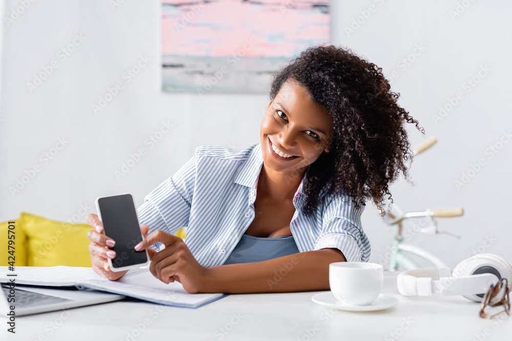 Cheerful african american teleworker holding smartphone with blank screen near laptop, headphones and cup on blurred foreground Cheerful african american teleworker holding smartphone with blank screen near laptop, headphones and cup on blurred foreground