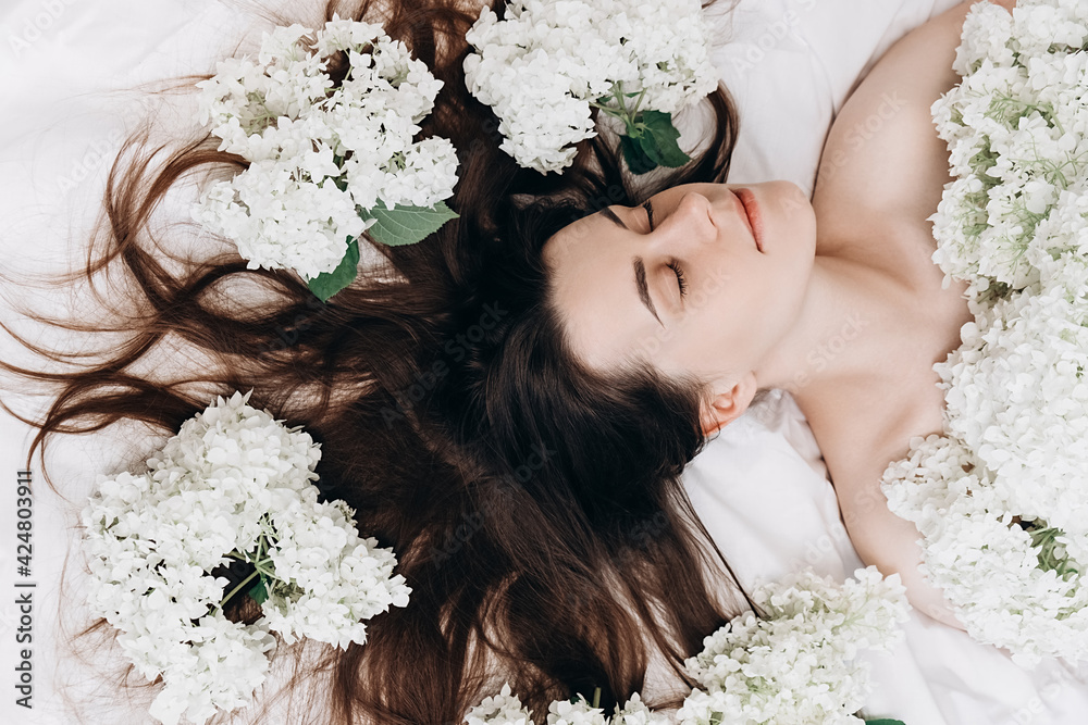Naklejka premium Top view beauty portrait of sexy brunette girl lying on comfy white bed near small beautiful flowers. Slender young woman, perfect clean skin and long hair. Natural cosmetics for skin care.