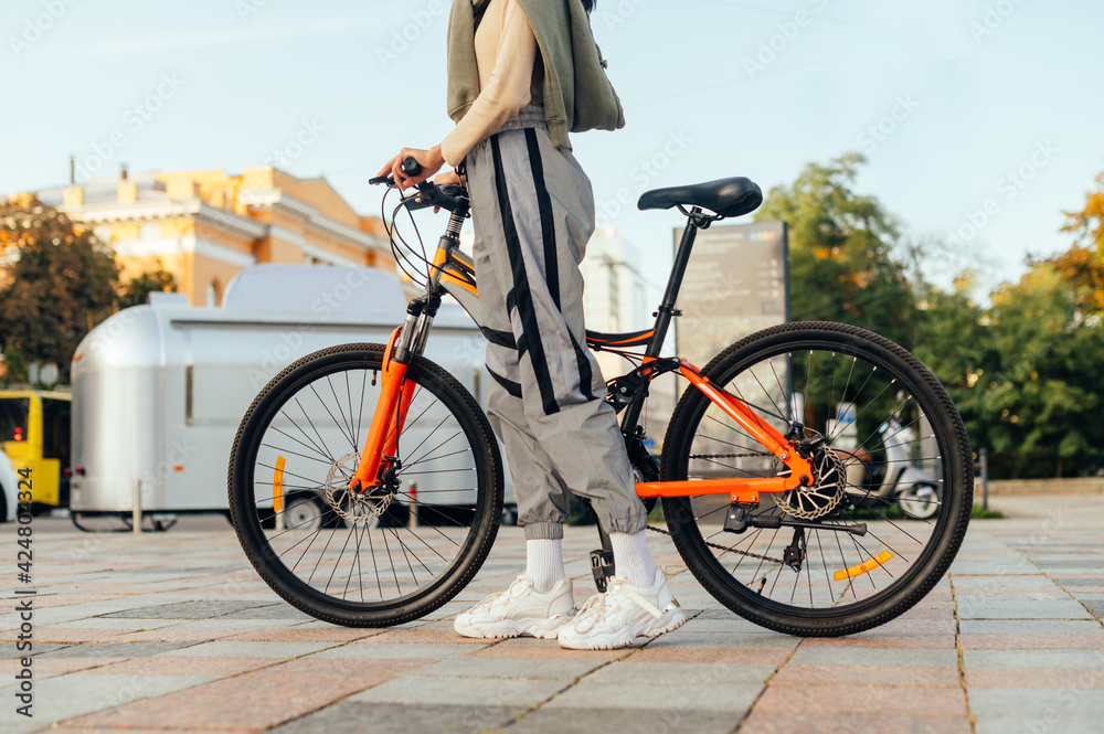 Cropped photo of a modern colorful bright orange bike and woman's legs in stylish pants and white training shoes in urban scenery. Details.